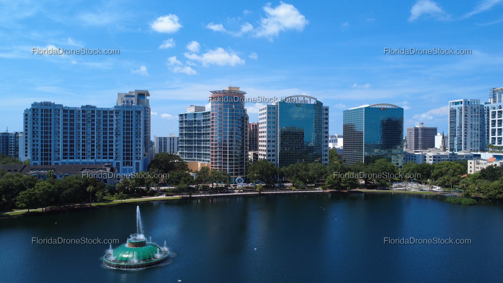 Clean summer aerial of the full downtown Orlando office-tower skyline with the Linton E. Allen Memorial Fountain in the foreground at Lake Eola Park.